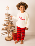 Toddler wearing the cream I Believe knitted jumper with red striped leggings beside a Christmas tree